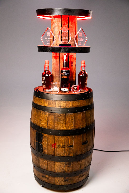 A rustic wooden barrel converted into a decorative display, featuring bottles of bourbon, glassware, and awards. The display is illuminated with red accent lighting and includes two circular shelves. The barrel is labeled with the Studio46 logo.